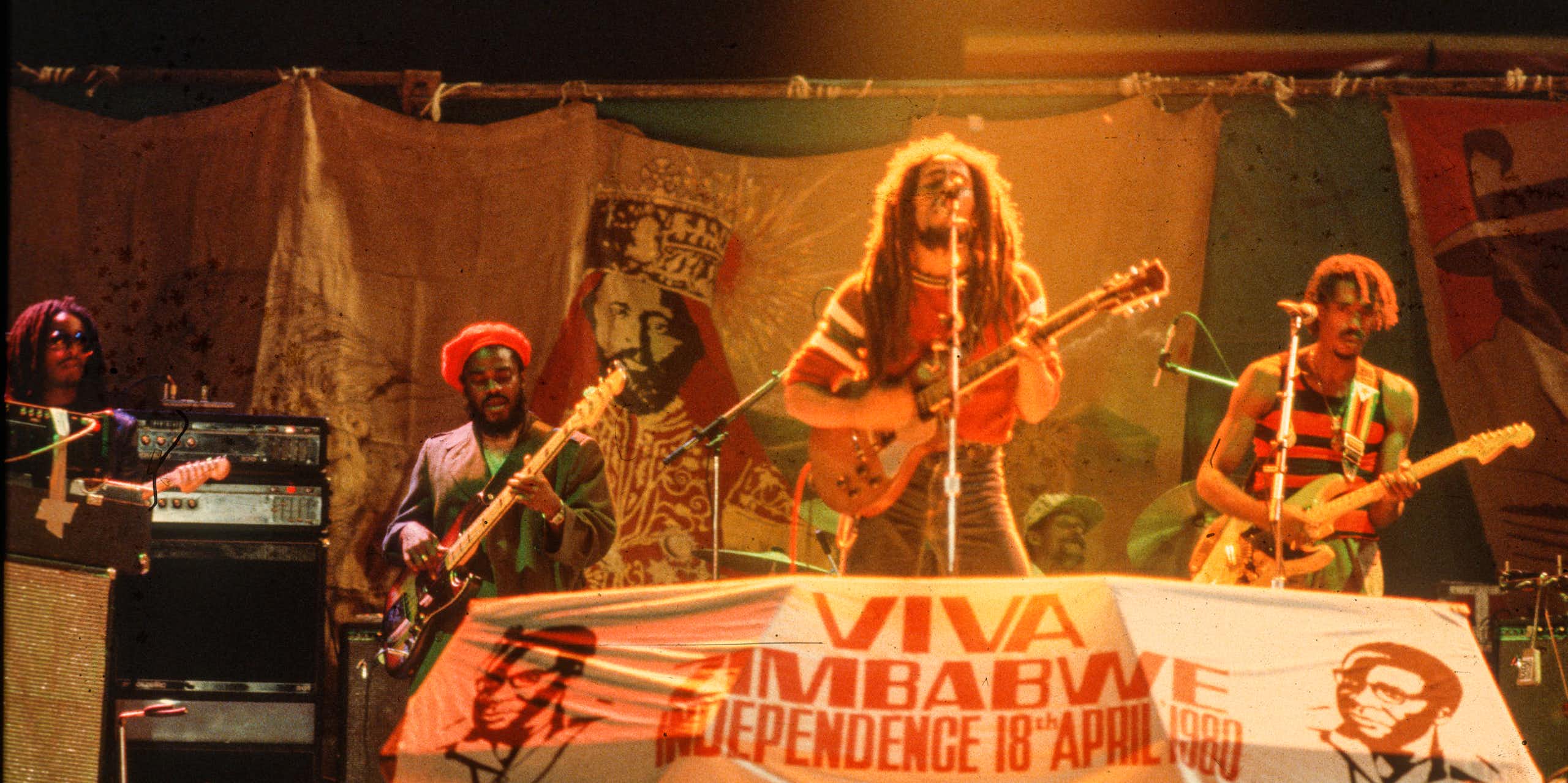 Dreadlocked man sings and plays guitar in front of sign reading 'Viva Zimbabwe.'