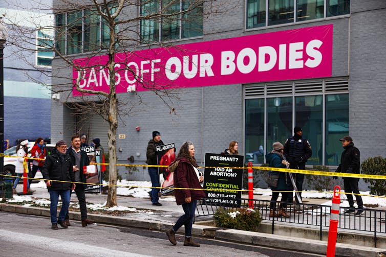 People, some holding posters, march outside of a grey building that says 'Bans off our bodies' in white writing, against a hot pink backdrop.
