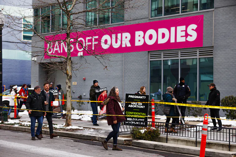 People, some holding posters, march outside of a grey building that says 'Bans off our bodies' in white writing, against a hot pink backdrop.