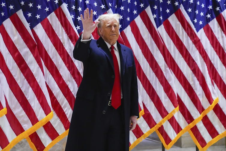A man in an overcoat waves while standing in front of a row of American flags.