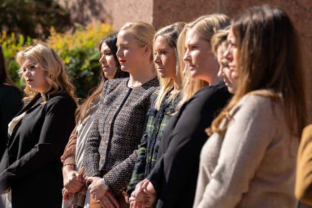 Seven women stand in a row and wear formal clothing, looking ahead. 