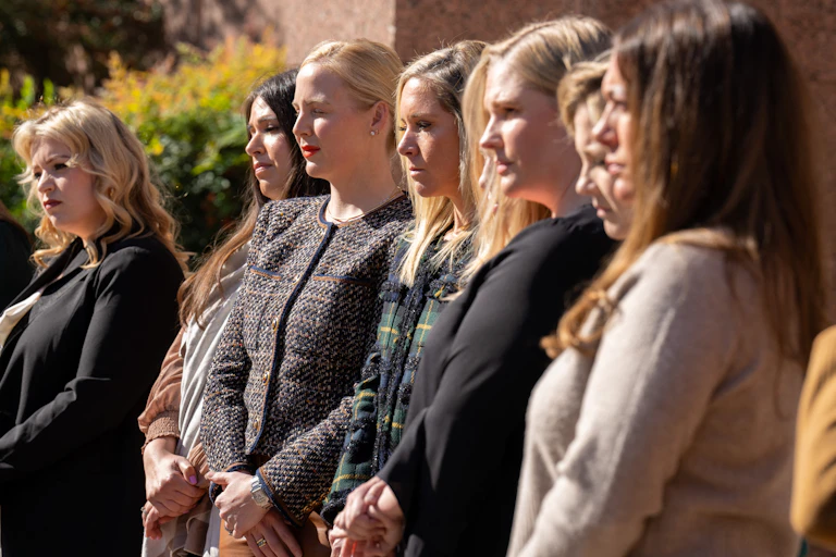 Seven women stand in a row and wear formal clothing, looking ahead.