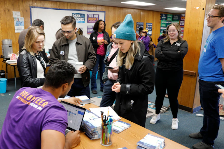 Group of young white people standing in panel walled room talking to man at desk with a computer.