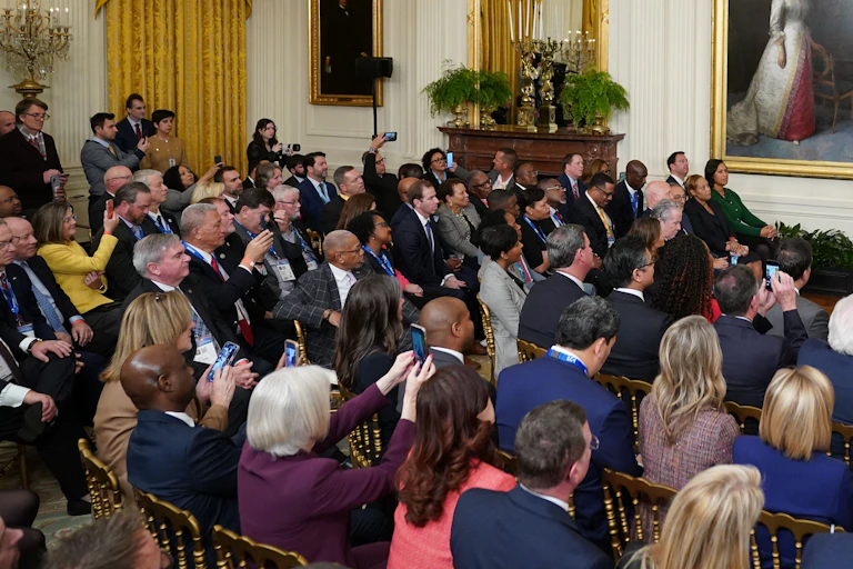 A man in a suit stands on a stage addressing a group.