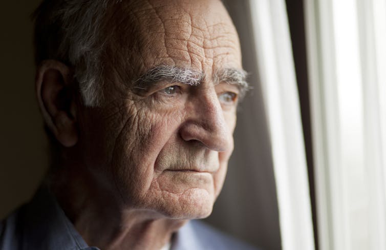 An older man with a serious expression on his face looks out a window.