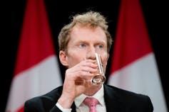 A man with dark blond hair sips on a glass of water.