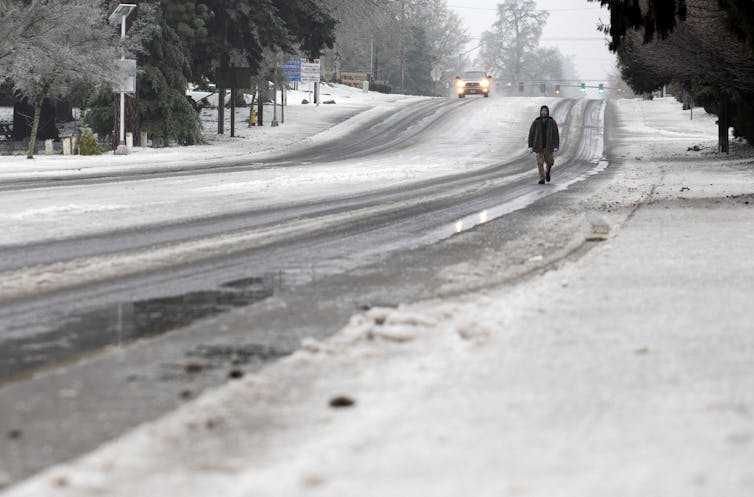 A person walks along a slushy road with a  car coming. Tree branches are coated in ice.