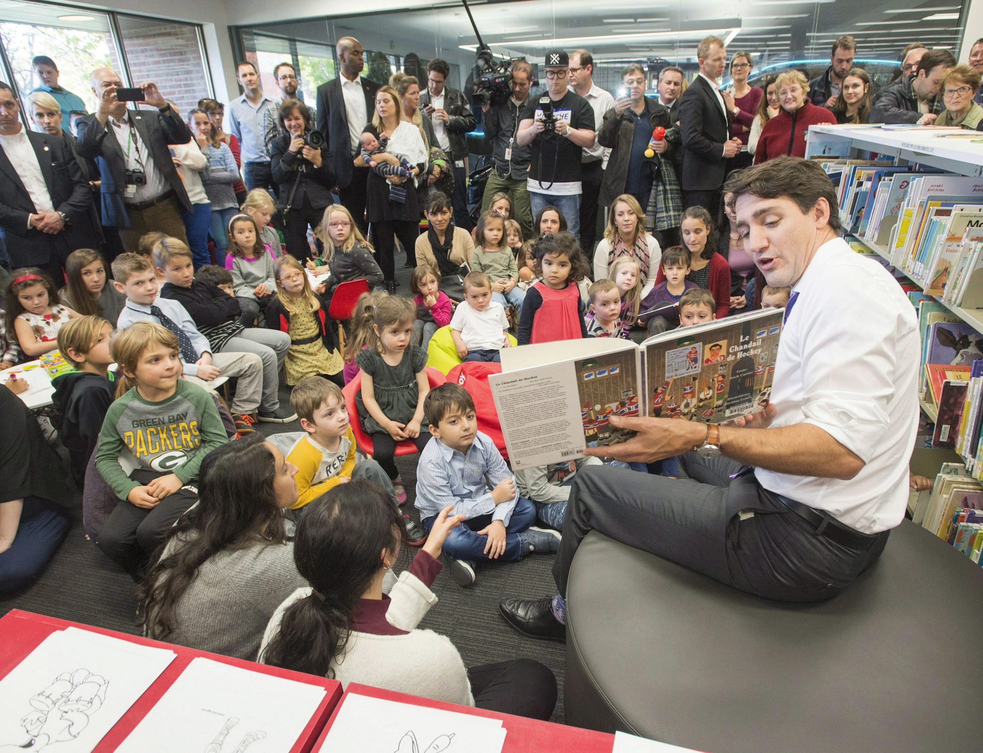 Justin Trudeau sits with his shirtsleeves rolled up, holding up a book, in front of a group of children