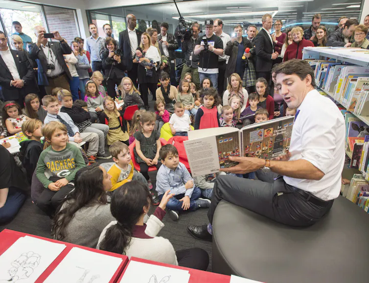 Justin Trudeau sits with his shirtsleeves rolled up, holding up a book, in front of a group of children