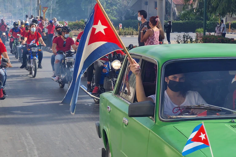 A man in a green car holding a Cuba flag aloft in front a line of motorcycles.