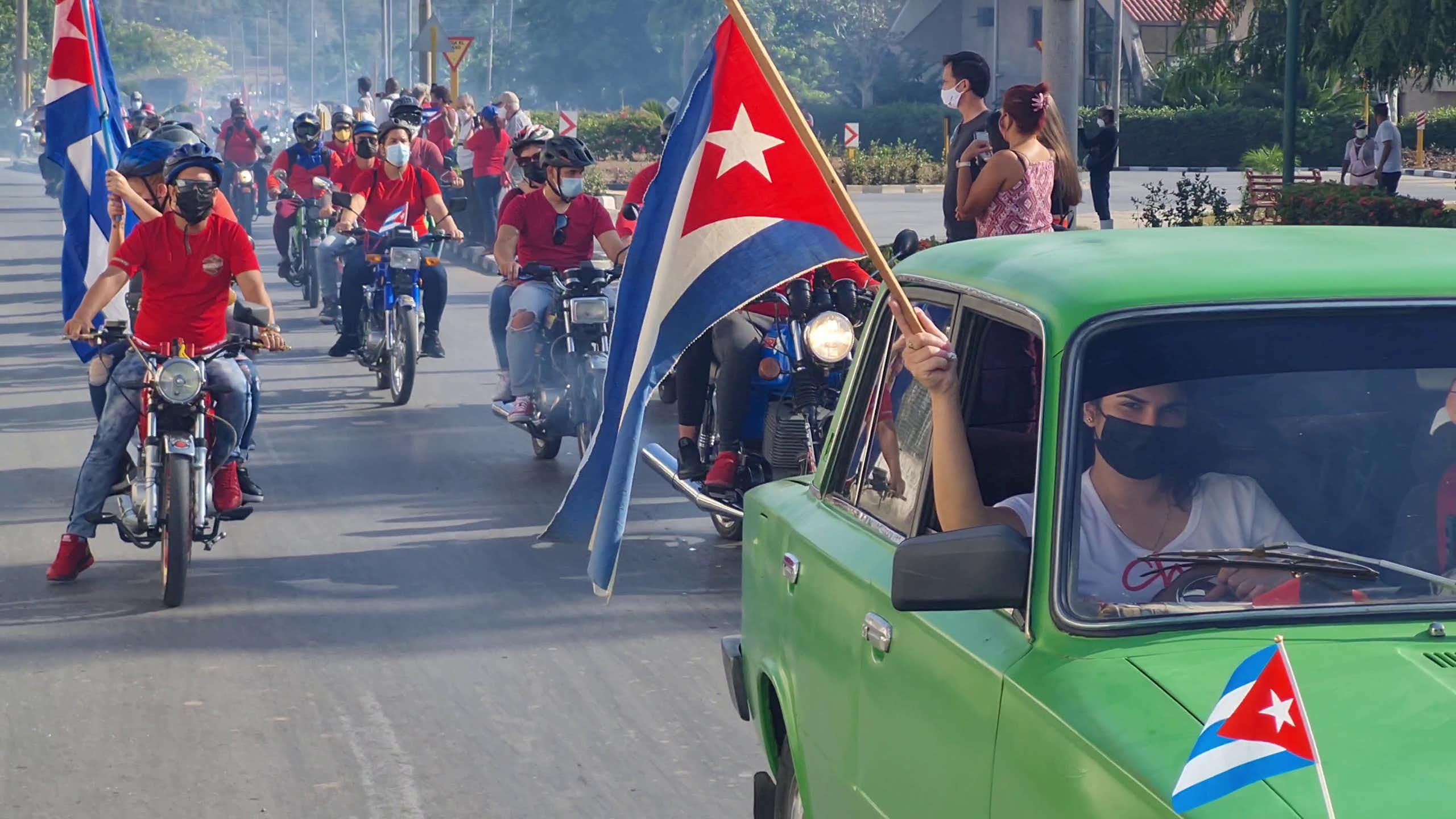A man in a green car holding a Cuba flag aloft in front a line of motorcycles.