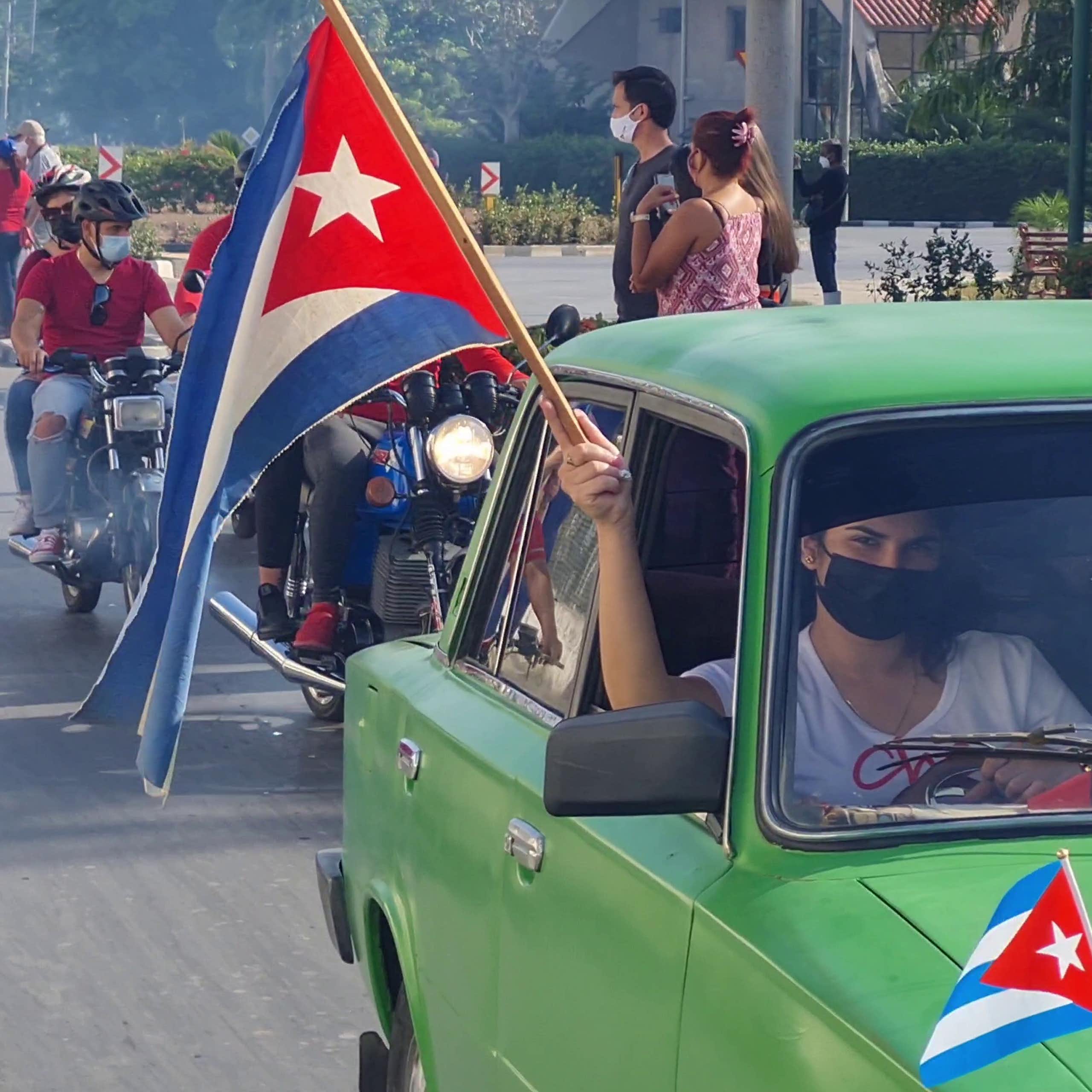 A man in a green car holding a Cuba flag aloft in front a line of motorcycles.