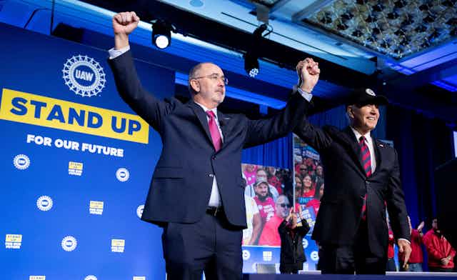 President Biden, wearing a baseball cap, holds hands aloft with a tall man with a close-shaven beard next to the UAW logo