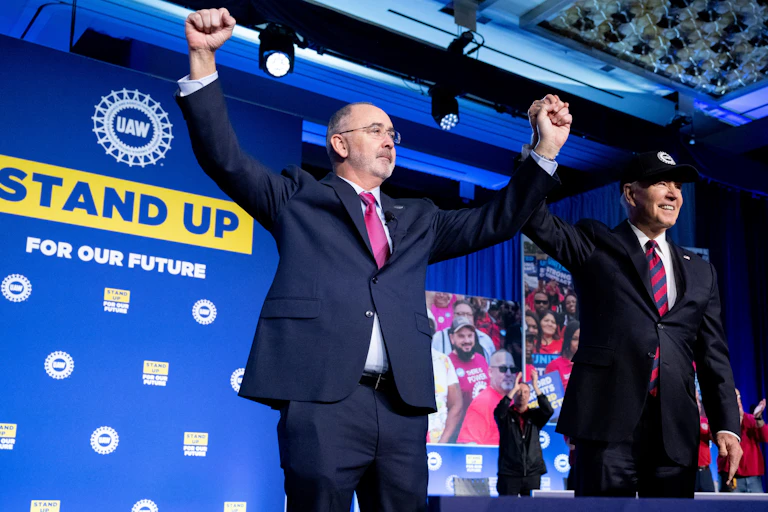 President Biden, wearing a baseball cap, holds hands aloft with a tall man with a close-shaven beard next to the UAW logo