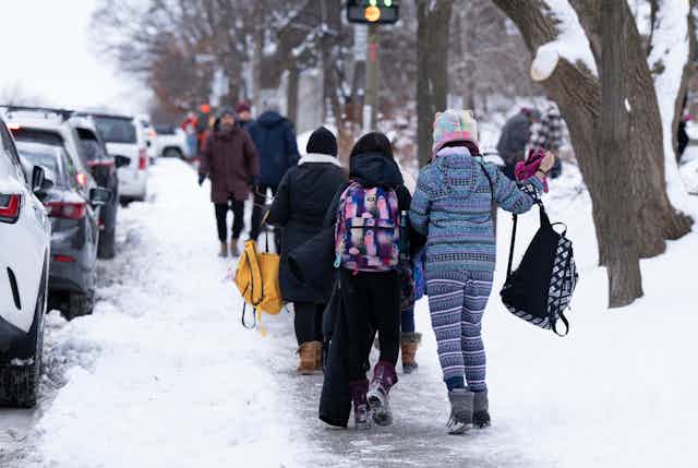 Students in winter coats and backpacks seen on a snowy sidewalk.