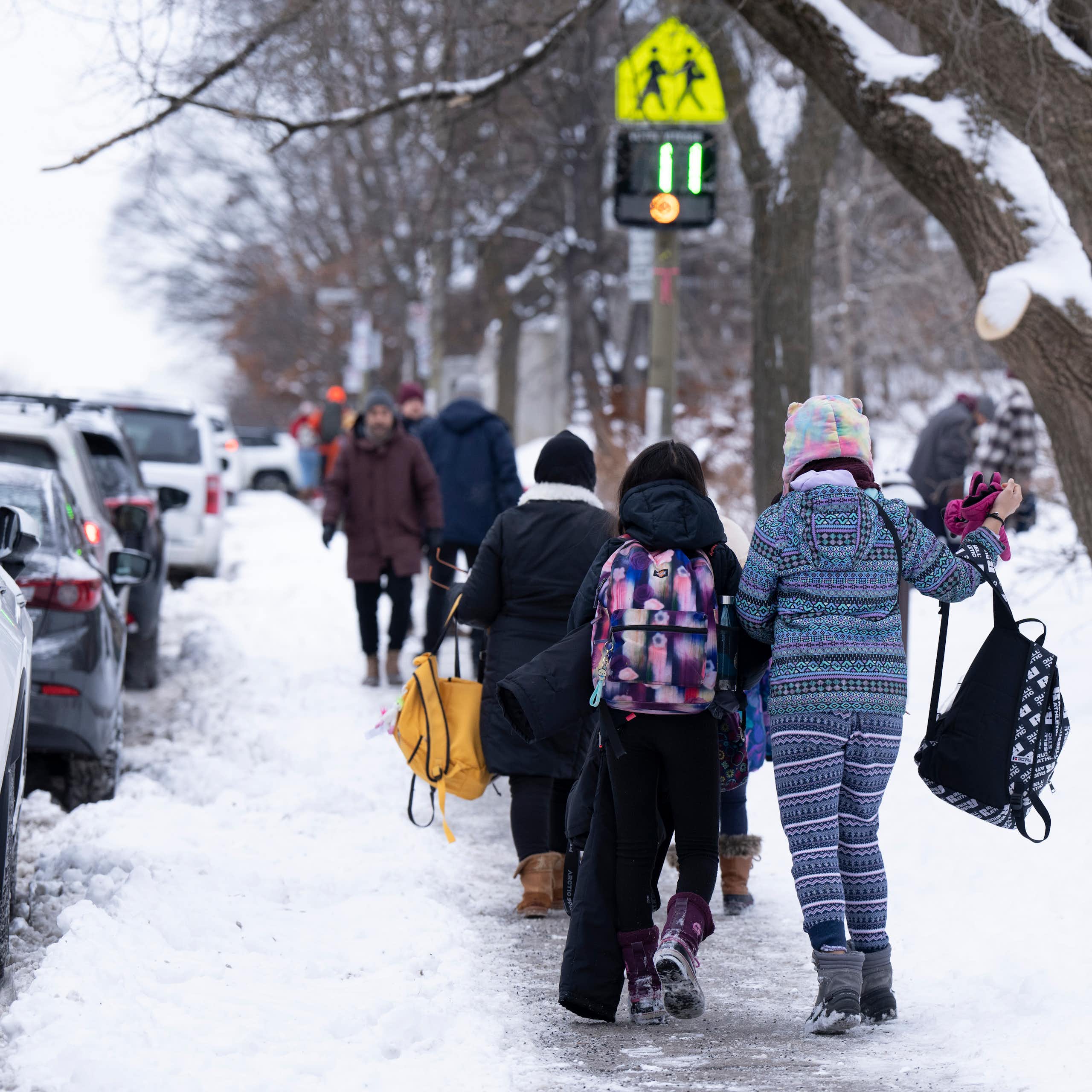 Students in winter coats and backpacks seen on a snowy sidewalk.