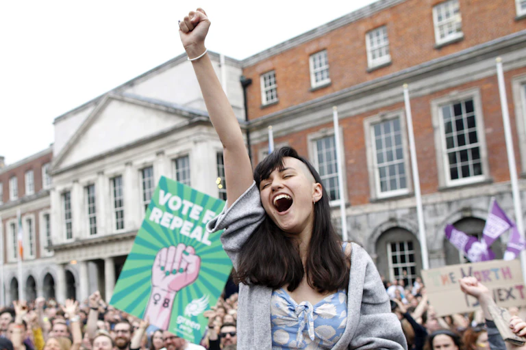 A woman on a man's shoulders cheers with her arm in the air at an outdoor gathering of mostly women.