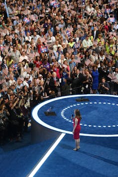 A large crowd gathers in front of a blue carpeted area where Chelsea Clinton is standing.