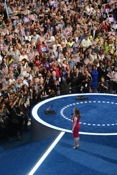A large crowd mass in front of a blue carpeted area where Chelsea Clinton stands.