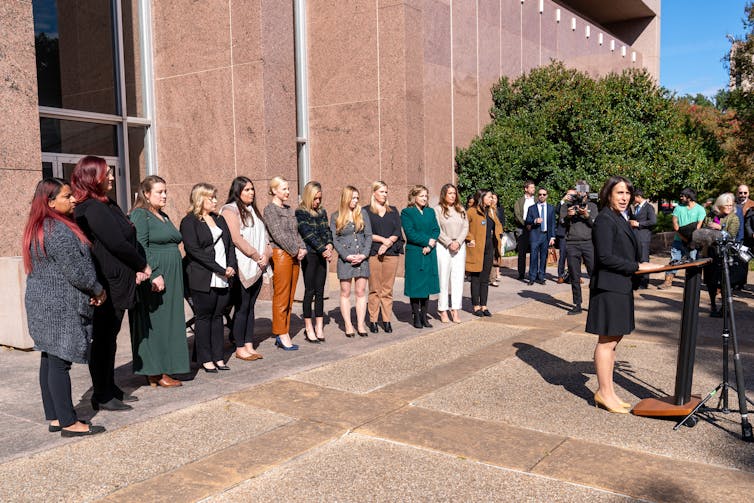 A woman in a black outfit stands at a podium in front of a long row of women who stand looking forward.