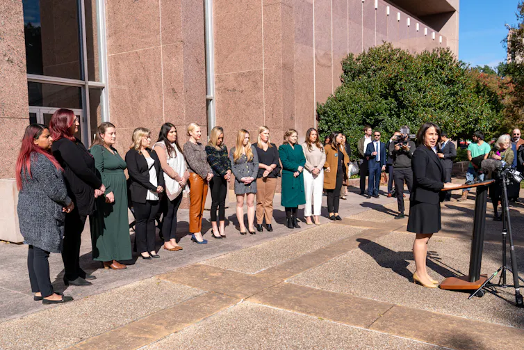 A woman in a black outfit stands at a podium in front of a long row of women who stand looking forward.