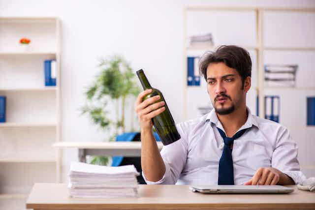 Un jeune homme en cravate est assis à la table de son bureau. Une de ses mains est posée sur son ordinateur portable fermé. De l'autre, il tient une bouteille de vin vide.
