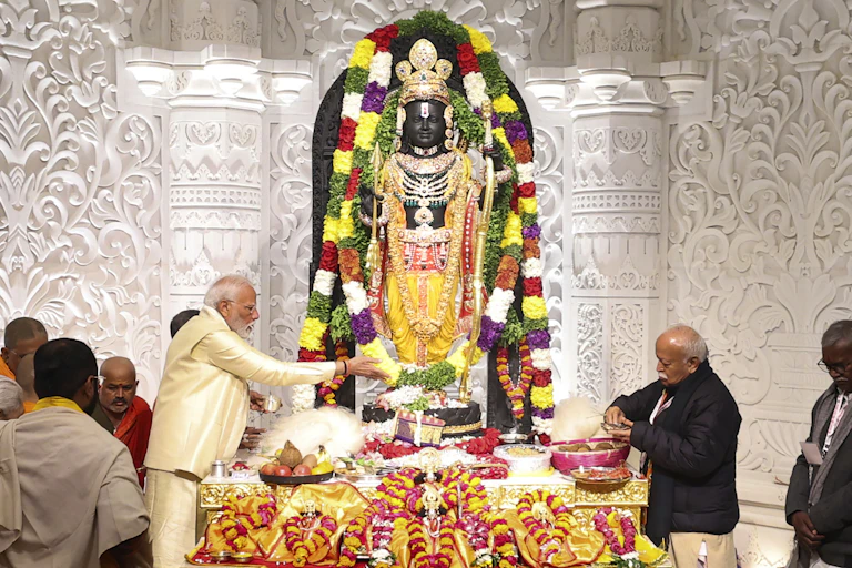 A garlanded standing icon of a deity in a temple, with two men, one in beige attire, performing rituals, while a few others stand near them.
