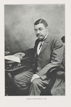 A Black man wearing a business suit sits at a desk with his right hand on a sheet of paper.