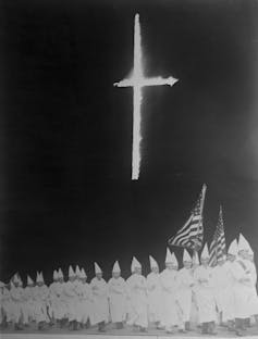 Underneath a burning cross, a group of white men dressed in white robes and white hoods march holding American flags.