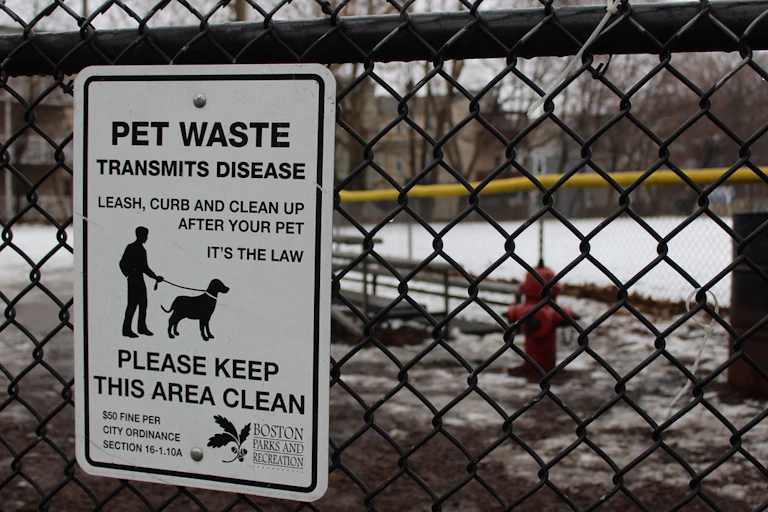 a sign on a fence warns 'Pet waste transmits disease' with dog park in background