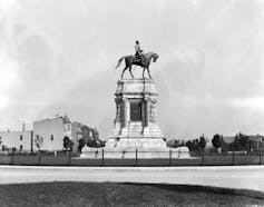 A large statue is seen in the middle of a park that depicts a white man siting atop a horse.