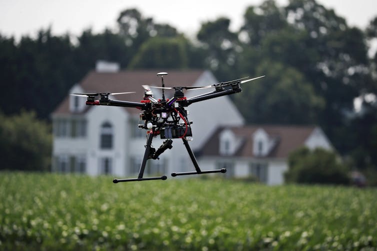 A small drone flying over a crop field, with a house in the background.