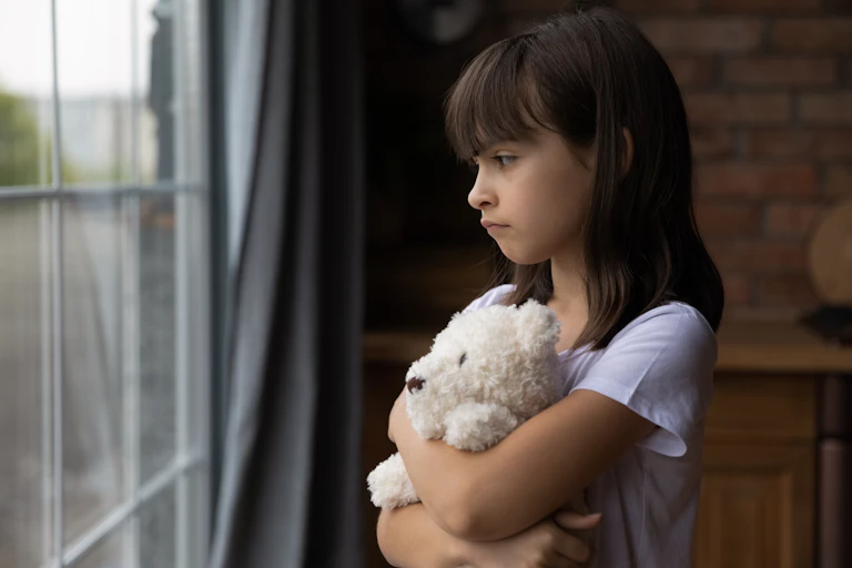 A child hugging a stuffed toy, looking out a window looking unhappy