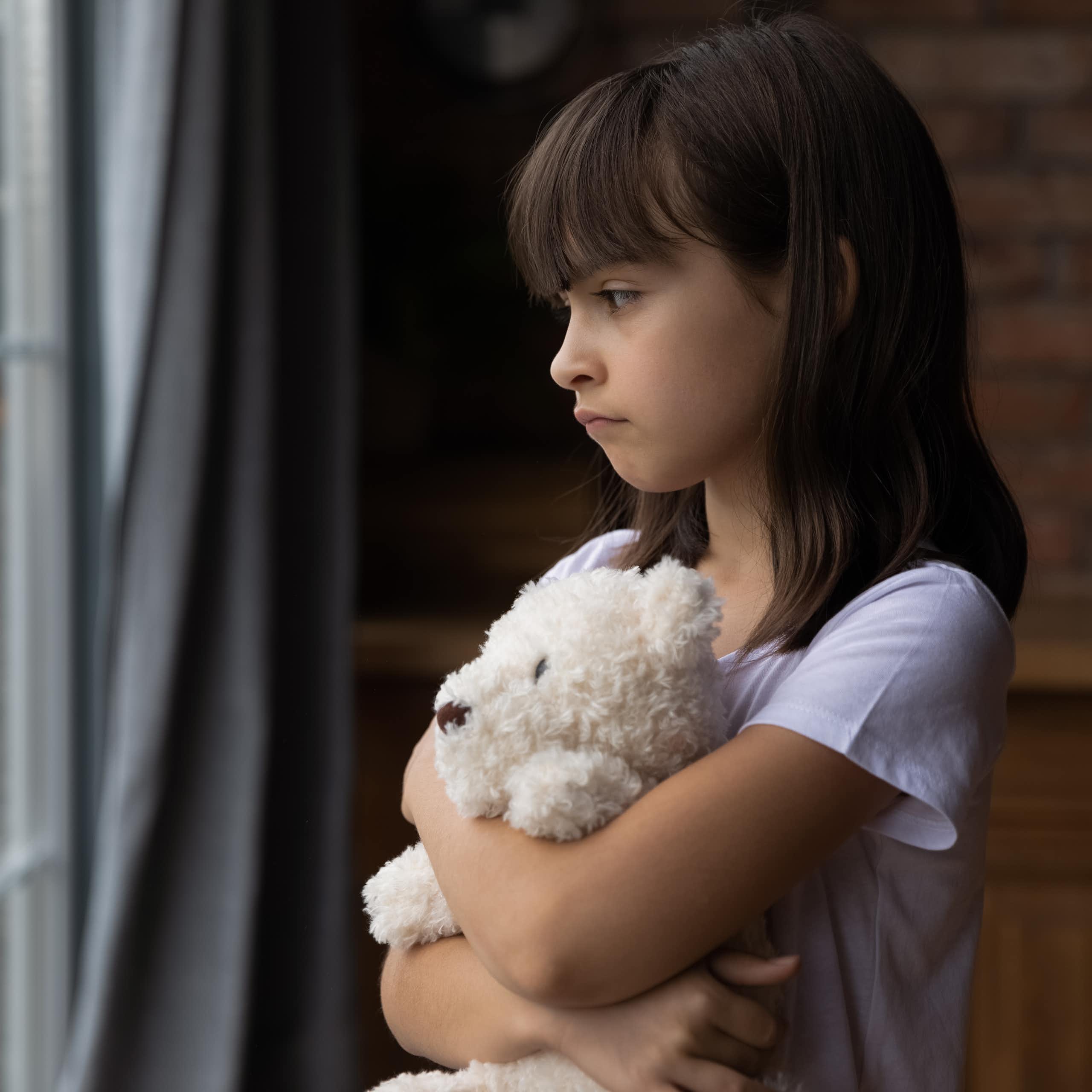A child hugging a stuffed toy, looking out a window looking unhappy