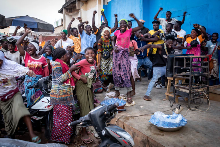 A crowd of people in the street cheer and raise their arms, beaming. At the centre of attention is a TV screen on a table propped against a metal shelf.