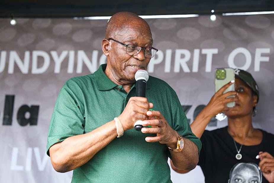 A bald man in glasses and a green golf shirt speaks into a microphone as a woman behind him holds her cellphone up , trained on him.