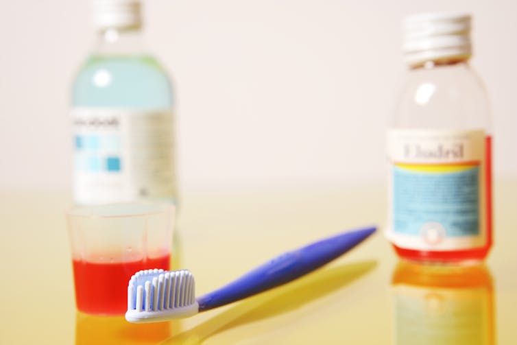 Two clear bottles with white screw tops, and illegible labels. One of the bottles is half full of red liquid and the other is full of a light blue liquid. A blue toothbrush is between the bottles, with a clear mouthwash cup containing red fluid.