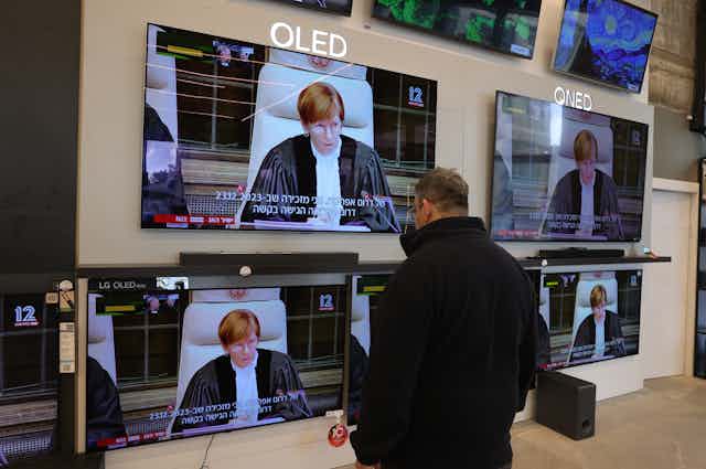 A man looks watches a bank of televisions throiugh a shop window. On the screen is a judge giving a ruling