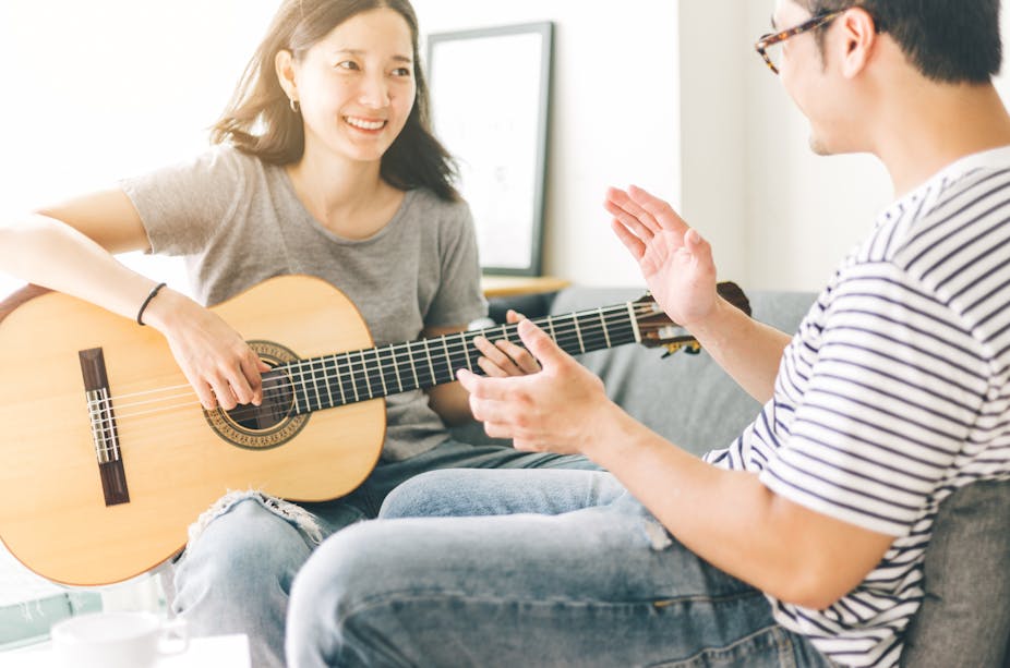 A young woman plays a guitar to a man sat next to her. 