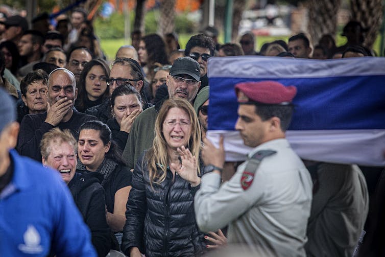 A soldier wearing a red beret carries a coffin covered in a blue and white cloth. People stand behind him crying.