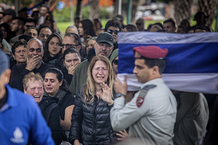 Un soldado con boina roja lleva un ataúd cubierto con una tela azul y blanca.  La gente llora detrás de él.