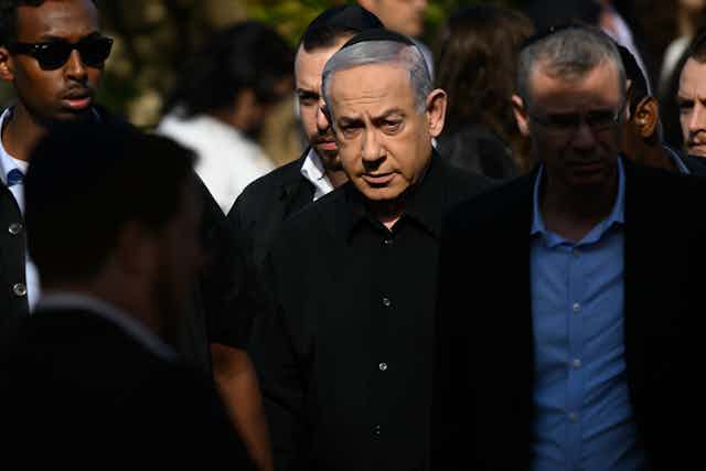 Benjamin Netanyahu wears a black button down shirt and a black kippah and stands in a group of other men. 