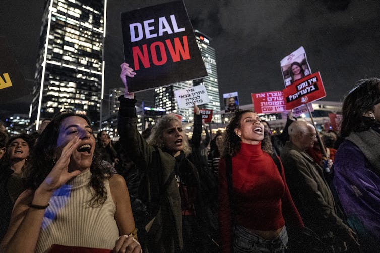 A group of people, including several women, hold signs and shout in a nighttime shot, in front of tall, lit up buildings. One of the signs says 'Deal now.'