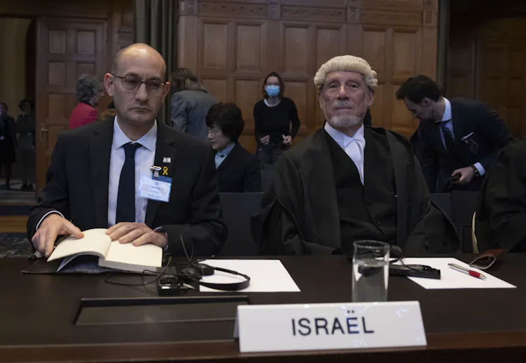 Two men wearing dark jackets - one wearing a tie and the other wearing an old fashioned white wig - sit together at a table with the words Israel in a placard at the front of the table.