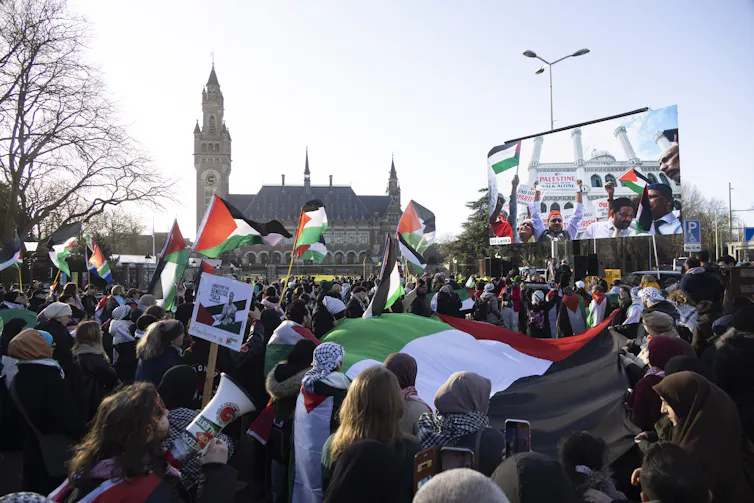 A large crowd of people wave Palestinian flags outside of a tall European building on a grey day.
