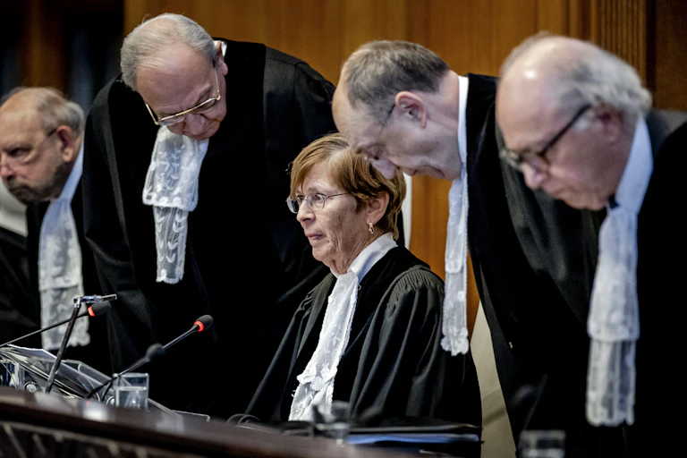 A woman in a black robe with a white scarf sits at a table, with four men dressed similarly stand nearby, with their heads bent forward.