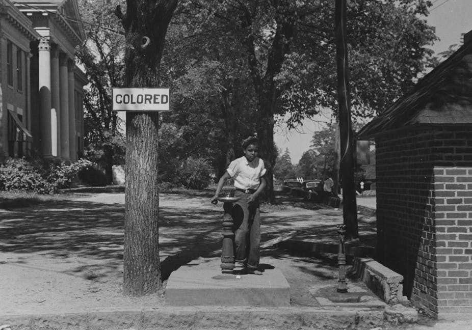 A Black girl is seen using  a water fountain near a sign that reads colored.