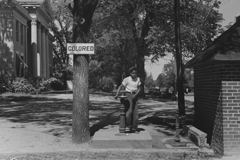 A Black girl is seen using a water fountain near a sign that reads colored.