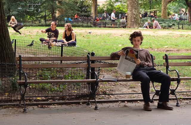 A young person sits on a bench in a well-used park reading the paper.