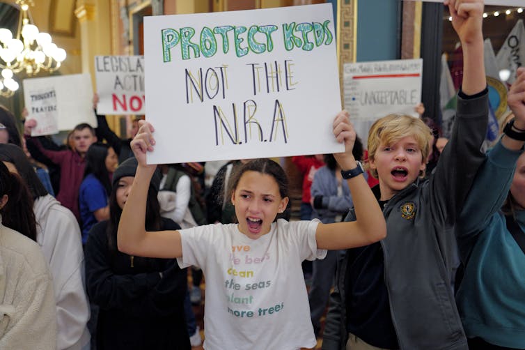 Kids speak out at a rally, with one holding a sign saying 'protect kids, not the NRA.'
